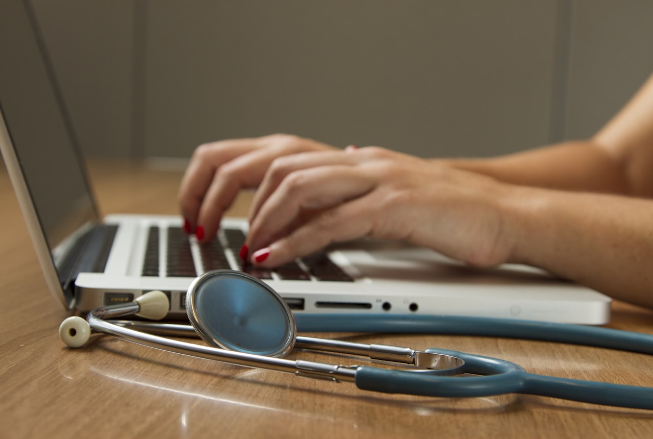 Hands typing on a laptop beside a stethoscope